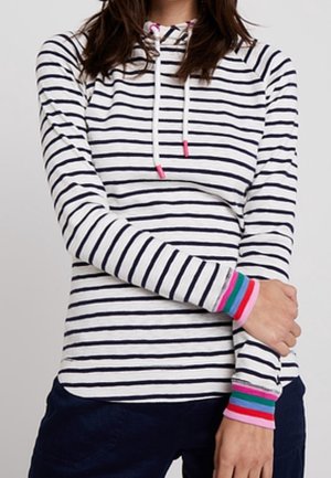 Frau trägt ein weißes Langarmshirt mit schwarzen horizontalen Streifen und bunten gestreiften Bündchen, kombiniert mit dunkler Hose.