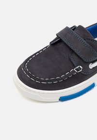 Scarpa di pelle blu scuro con cuciture bianche, cinturino in velcro e suola in gomma blu texturizzata, con un design casual da scarpa da barca.
