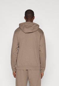 Hnedá mikina s kapucňou, vo voľnom strihu, vyrobená z mäkkej tkaniny. Obsahuje klokanie vrecko a rebrované manžety a spodný lem.