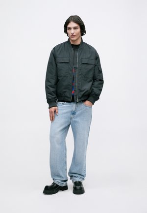 Sorte bomberjakke med to forlommer over en mønstret sweater, parret med lyseblå, løst siddende jeans og sorte sko med tykke såler.