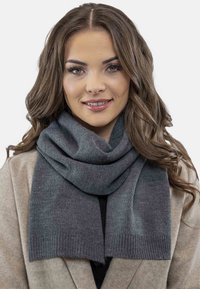 Écharpe grise en tricot doux, de forme rectangulaire avec des bords côtelés, portée lâche autour du cou sur un manteau beige.