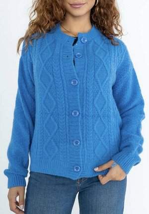 Femme portant un cardigan bleu en tricot torsadé boutonné et un jean bleu, avec une main dans la poche et des cheveux bouclés visibles.