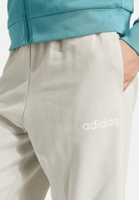 Light gray sweatpants with a smooth texture and loose fit. Features a white "adidas" logo on the left thigh. Accompanied by a blue upper.