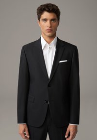 Giacca nera con due bottoni, rever a tromba e tasche frontali. Camicia bianca sotto, con colletto semplice e un fazzoletto nella tasca.