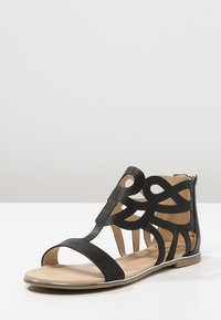 Svart lädersandal med utskärningsdesign, öppen tå, ankellängd och platt sula. Har eleganta remmar och en tanfärgad innersula.