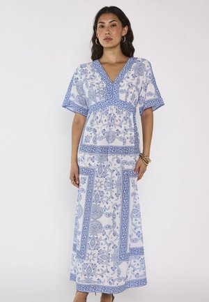Donna che indossa un vestito maxi bianco e blu a fantasia con scollo a V e maniche corte, con orecchini a cerchio dorati e bracciali su sfondo uniforme.