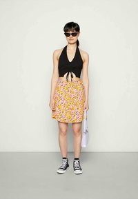 Mulher vestindo um top halter preto, uma saia floral amarela e rosa, tênis altos pretos, óculos escuros pretos, segurando uma pequena bolsa branca.