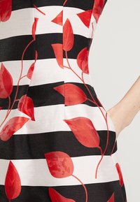 Mustavalkoinen vaakaraidoitettu mekko, jossa on punaisia lehtikuvioita ja k�äsi oikeassa taskussa vaaleaa taustaa vasten.