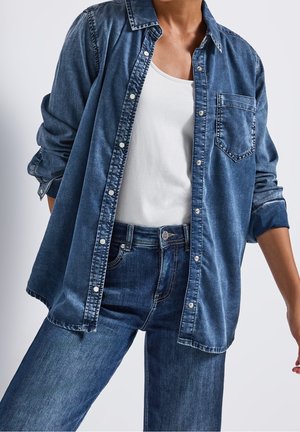 Persoon draagt een open blauwe spijkerblouse over een wit hemdje met een bijpassende blauwe spijkerbroek tegen een effen achtergrond.
