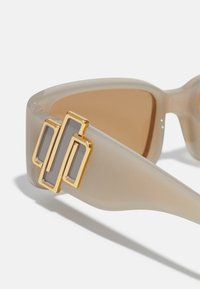 Gafas de sol en plástico translúcido beige con lentes marrones, con un acento geométrico dorado en el costado. Textura suave y diseño angular.