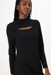 Robe noire à manches longues en tissu lisse, avec un col montant et un découpage au niveau de la poitrine pour un intérêt visuel.