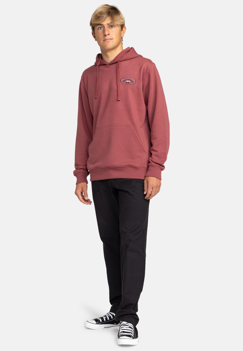 Sudadera granate con un bolsillo frontal, capucha ajustable con cordones y logo bordado. Combinada con pantalones negros y zapatillas negras.