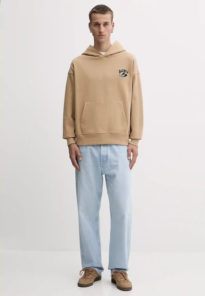 Sudadera beige con un bolsillo frontal y detalle del logo, combinada con jeans de pierna recta azul claro y deportivas marrones con cordones.