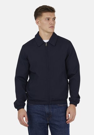 Młody mężczyzna w ciemnoniebieskiej kurtce zapinanej na zamek błyskawiczny z elastycznymi mankietami i kołnierzem, w połączeniu z niebieskimi dżinsami, patrzący na bok.