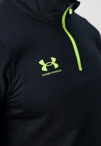 Svart halvdraget pullover i texturerat tyg, med en neongrön dragkedja och logotyp med texten "UNDER ARMOUR" på bröstområdet.