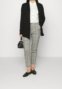 Zwarte lange cardigan, witte blouse met knopen, grijze geruite broek, zwarte platte schoenen en een kleine zwarte handtas. Outfit benadrukt neutrale tinten en patronen.
