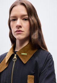 Νεαρή γυναίκα με μακριά καστανά μαλλιά, που φοράει ένα μπλε σκούρο μπουφάν με καφέ γιακά από κορδόνι και ένα πράσινο καρφίτσα "SPEY" κοντά στο φερμουάρ.