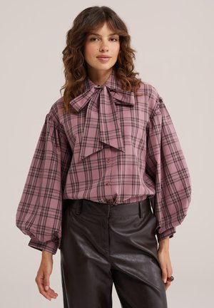 Femme portant un chemisier à carreaux rose avec un grand nœud papillon et des manches bouffantes, associé à un pantalon en cuir noir sur un fond uni.