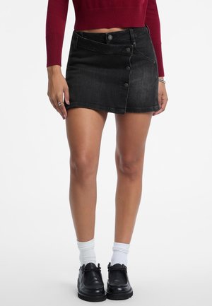 Schwarzer Denim-Rock mit taillierter Form, ausgestattet mit einem seitlichen Knopfverschluss und einem rohen Saum. Kombiniert mit schwarzen Schuhen und weißen Socken.
