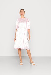 Vestido de cetim rosa claro com mangas bufantes, detalhes em renda e uma cintura ajustada. Combinado com sandálias de salto bloco bege e uma pequena carteira.