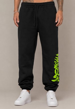 Uomo con pantaloni della tuta neri con scritta graffiti verde sulla gamba sinistra e sneakers bianche, in piedi davanti a uno sfondo neutro.