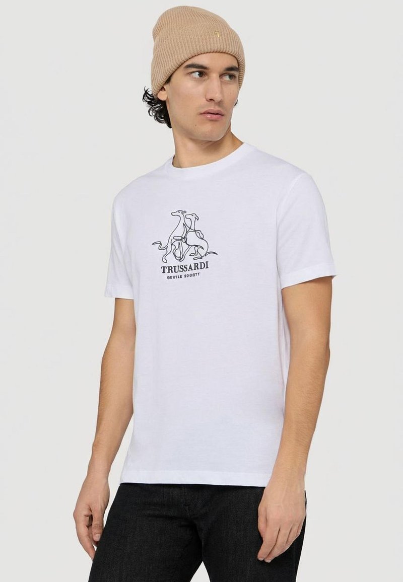 Camiseta blanca de algodón con un gráfico negro de perros y el texto "TRUSSARDI" debajo. Combina con un gorro de punto acanalado beige.