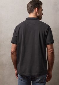 Homme aux cheveux courts et foncés portant un polo noir à manches courtes et un jean bleu, debout contre un mur gris clair uni, de dos.