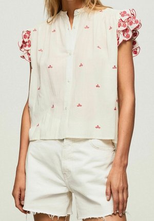 Kvinde iført hvid ærmeløs knapbluse med rød blomsterbroderi og flæseærmer, kombineret med hvide denimshorts.