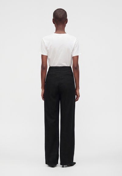Personne debout de dos portant une chemise blanche à manches courtes et un pantalon noir taille haute à jambes larges avec des talons sur un fond uni.