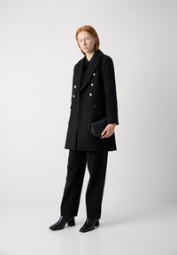 Abrigo negro de doble botonadura con botones en tono plateado, llevado sobre una blusa negra y combinado con pantalones negros y botas de tacón cuadrado. Clutch negro.