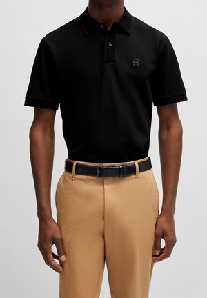 Mann trägt schwarzes kurzärmeliges Poloshirt und beige Hose mit schwarzem Gürtel, von Hals bis Hüfte vor einfarbigem Hintergrund gezeigt.