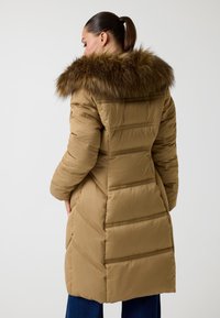 Cappotto lungo imbottito marrone con cappuccio bordato in pelliccia, caratterizzato da un design a zig zag trapuntato e una texture liscia, visto da dietro.