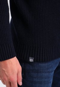 Pull en maille bleu marine avec texture côtelée, doté d'une petite étiquette logo sur l'ourlet. Porté avec un jean denim bleu.