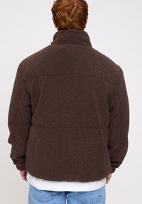 Brown fleece jacket with a high collar, textured material, relaxed fit, and two seams on the back. Worn over a white shirt and blue jeans.