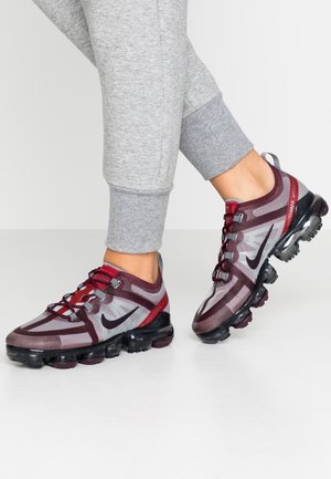 Personne portant un pantalon de survêtement gris et des baskets Nike Air VaporMax bordeaux et noires, debout sur une surface blanche.