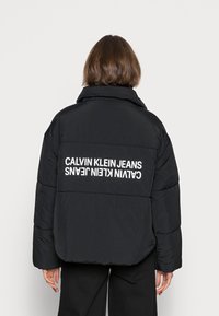 Svart puffjacka med hög krage och oversized passform. Visar en vit "CALVIN KLEIN JEANS" logotyp tydligt på baksidan.