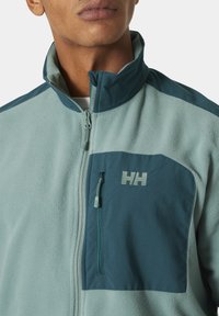 Chaqueta de forro polar en verde claro y azul oscuro, con un cuello alto, bolsillo en el pecho con cremallera y detalle del logo. Textura suave con paneles contrastantes.