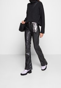 Schwarzer gerippter Rollkragenpullover, hochgeschnittene, glänzende schwarze Kunstlederhosen und pastelllila Plateauschuhe mit einer kleinen schwarzen Tasche und einem Seidenschal.