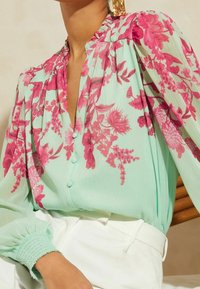 Blusa verde menta clara con patrón floral en rosa, escote fruncido y mangas abullonadas. Presenta cierre de botones y puños texturizados.