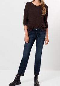 Haut marron foncé à manches longues associé à un jean bleu foncé ajusté. Le jean présente une coupe classique, avec des revers relevés, complété par des bottines noires.