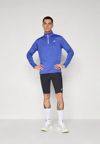 Blue long-sleeve athletic top with a quarter zip, paired with black cycling shorts. Model wears white athletic shoes and knee-high socks.