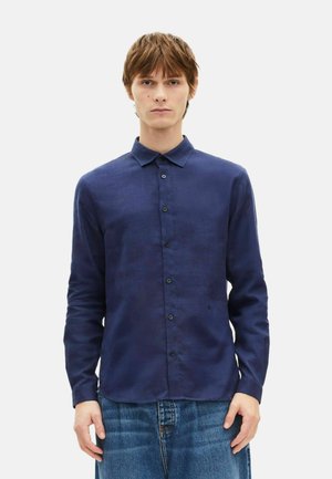 Jeune homme debout, portant une chemise à manches longues bleu marine boutonnée et un jean bleu, sur un fond clair uni.