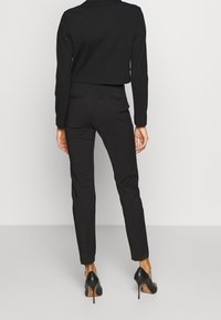 Femme vue de dos, portant un pantalon noir ajusté, une veste noire courte et des chaussures à talons hauts noires, sur un fond blanc.