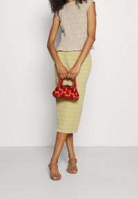 Top beige arricciato, gonna midi a coste verde chiaro e borsa intrecciata rossa con manico a treccia. Sandali con tacco a blocco trasparente completano il look.