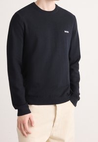 Homme portant un pull noir à manches longues avec le logo « BOSS » sur la poitrine et un pantalon beige clair, debout avec la main gauche dans la poche.