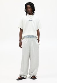White oversized t-shirt with navy embroidered text, paired with light grey baggy sweatpants and black slide sandals. Notable relaxed fit.