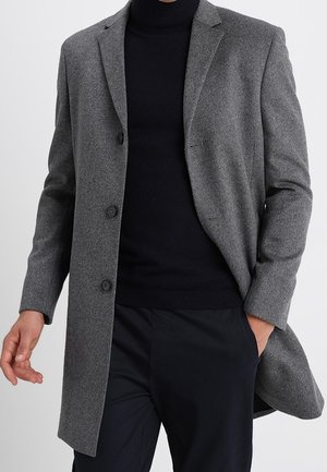 Muž nosící šedý kabát střední délky přes černý rolák a černé kalhoty, jedna ruka v kapse, druhá mírně natažená.