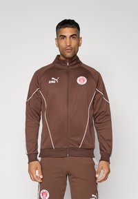 Chaqueta deportiva marrón con cuello alto, detalles en blanco y parches de logo, con cierre de cremallera y puños acanalados.