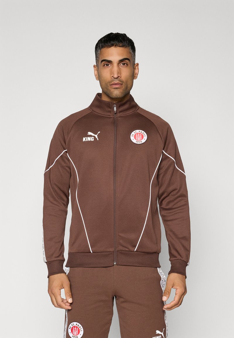 Chaqueta deportiva marrón con cuello alto, detalles en blanco y parches de logo, con cierre de cremallera y puños acanalados.