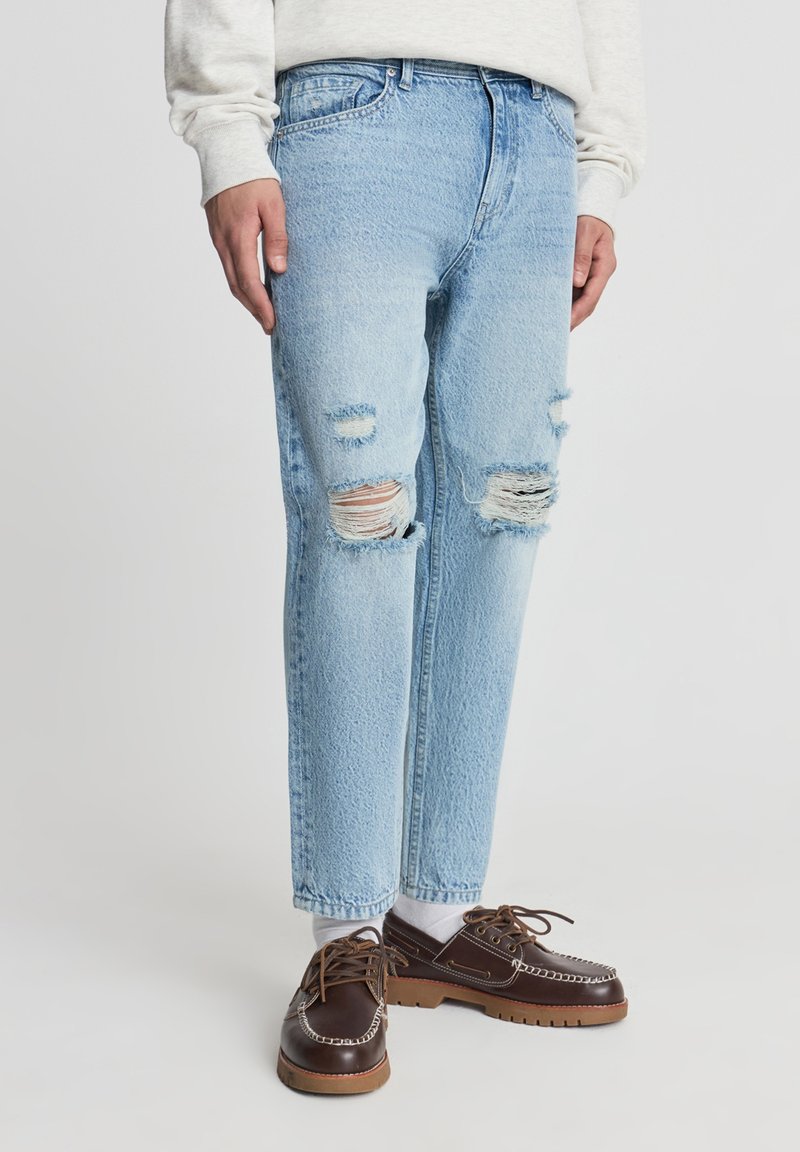 Jeans in denim azzurro chiaro con dettagli consumati e bordi sfrangiati sulle ginocchia. Indossati con scarpe marroni e calzini bianchi. Vestibilità comoda.